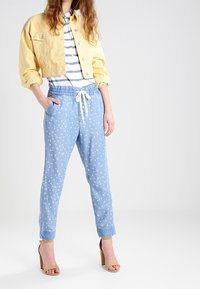 Gelbe, verkürzte Jeansjacke mit Knöpfen, gestreiftes T-Shirt in Weiß und Blau sowie blaue Hosen mit Zugband und weißem Blumenmuster, kombiniert mit beigen High Heels.