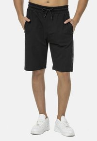 Schwarze Sportshorts mit elastischem Bund, Kordelzug, Seitentaschen und glatter Textur; kombiniert mit weißen Sneakers für einen Kontrast.