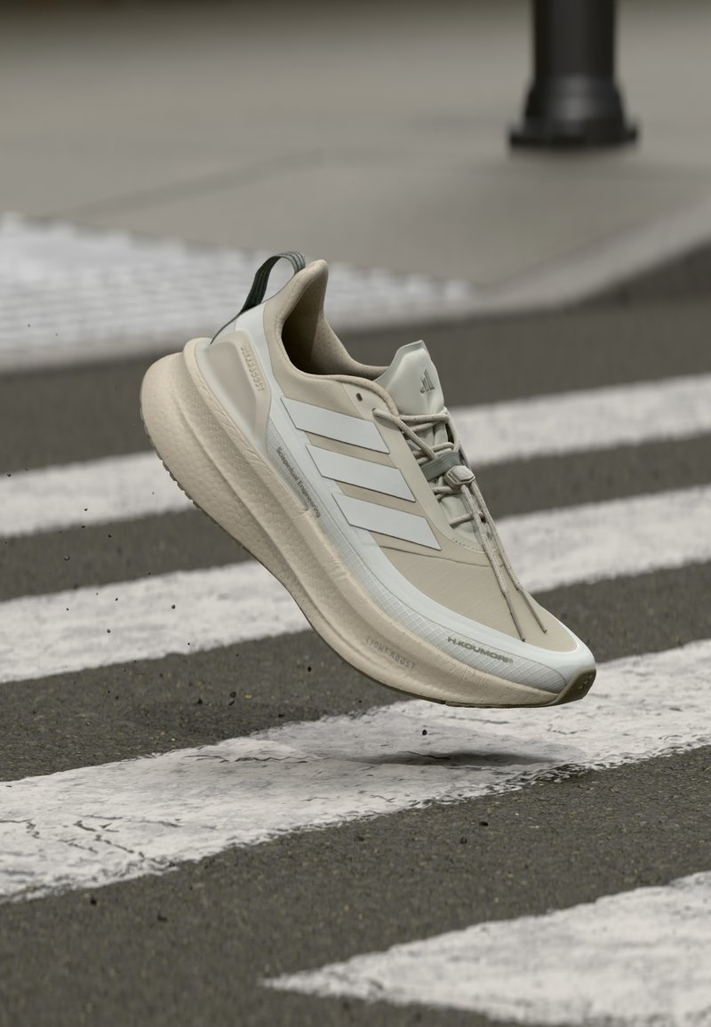 Pantof de alergare Adidas bej și alb, în aer, deasupra unei treceri de pietoni pe asfalt, evidențiind caracteristicile de greutate redusă și talpa boost.