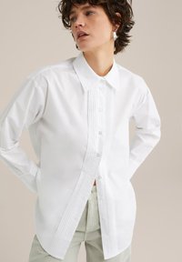 Biela košeľa s dlhým rukávom z bavlny so štruktúrovaným golierom, prednými záhybmi a zapínaním na gombíky, nosená s svetlozelenými nohavicami.