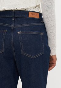 Dark blue denim jeans with a leather logo patch on the waistband, visible stitching, and two back pockets with a triangular design.