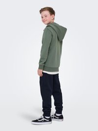 Felpa verde khaki con zipper frontale, vestibilità relaxed, e cappuccio regolabile, abbinata a pantaloni della tuta blu navy e sneaker nere.