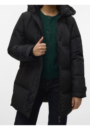 Personne portant une doudoune à capuche noire sur un pull vert boutonné et un pantalon sombre sur un fond uni.