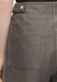 Gros plan d'une personne portant un pantalon couleur taupe avec une grande poche arrière carrée et une patte de taille boutonnée.