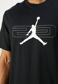 Sort T-shirt i bomuld med rund halsudskæring, prydet med en hvid grafik af en springende figur og tallet "23" i grå bagved.