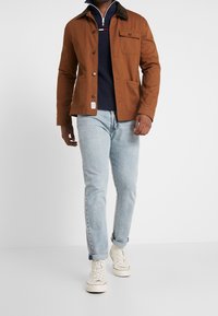 Brązowa bawełniana kurtka z kieszenią na piersi i zapięciem na guziki, zestawiona z granatowym swetrem z dzianiny, jasnoniebieskimi dżinsami i białymi trampkami.