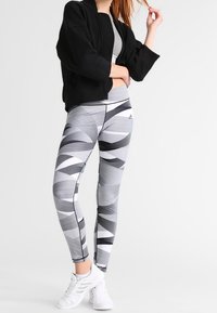 Chaqueta negra corta combinada con leggings grises y negros con un patrón de diseños geométricos. Zapatillas atléticas blancas completan el conjunto.