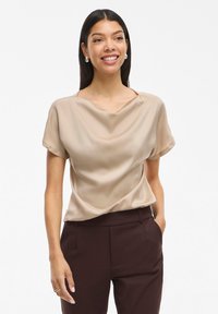 Blouse en satin beige avec un décolleté drapé et des manches courtes, associée à un pantalon sur mesure marron foncé. Texture lisse, design ajusté.