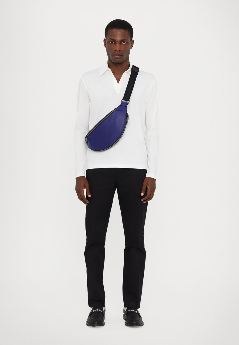 Zila ādas crossbody soma ar izliekta formu, melnu siksnu un rāvējslēdzēju, valkāta pāri baltai garām piedurknēm kreklam un melnām biksēm.