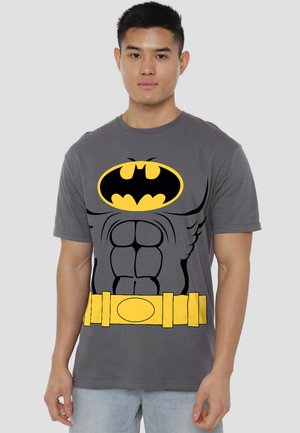 Young man wearing gray Batman-themed t-shirt with printed muscles, Batman logo, and yellow utility belt, standing against plain background.