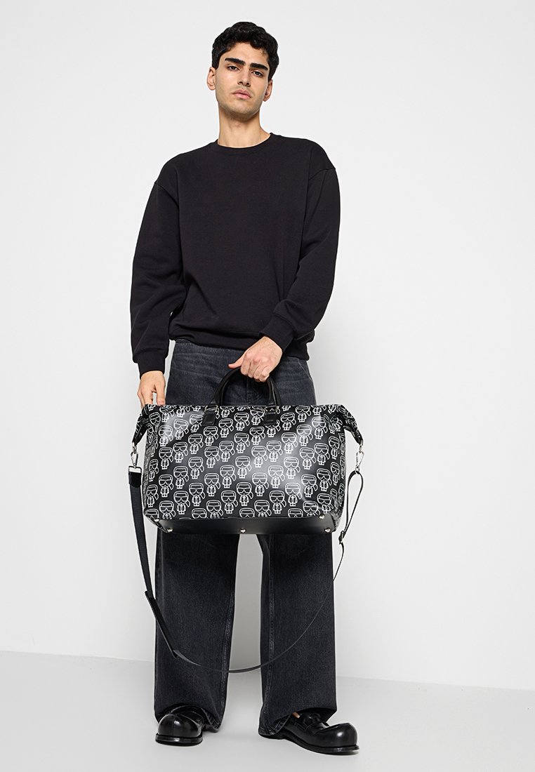 Bolsa de viaje negra con un patrón repetido de calaveras blancas, doble asa y una correa de hombro ajustable. Material liso con acabado brillante.