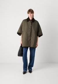 Geverfde olijfgroene jasje met een donkerbruin kraag, voorzien van een knoopsluiting en opgestikte zakken, gecombineerd met blauwe jeans en een zwarte handtas.