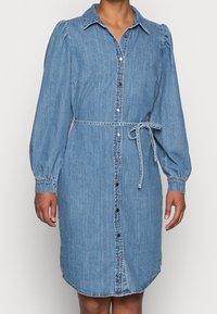 Jeansskjortklänning i ljusblått med långa puffärmar, knappförslutning fram, krage och midjeband. Har en böjd nederkant och sömnadsdetaljer.