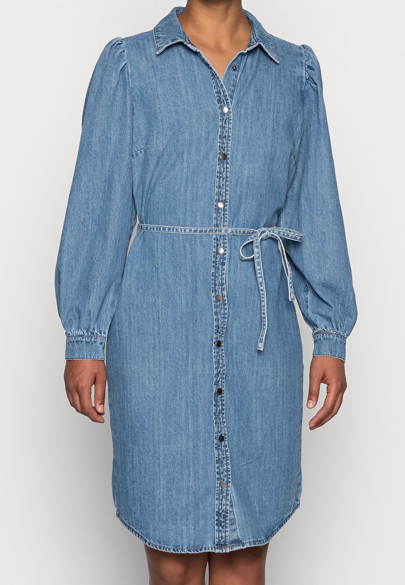 Jeansskjortklänning i ljusblått med långa puffärmar, knappförslutning fram, krage och midjeband. Har en böjd nederkant och sömnadsdetaljer.