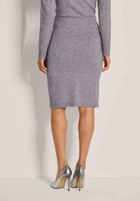 Frau in einem enganliegenden grauen Strickrock und passendem Oberteil, stehend vor einem schlichten hellen Hintergrund, sichtbar sind silberne High-Heels.