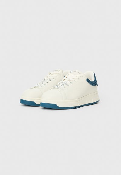Vita sneakers med en slät läderöverdel, blå gummisula och kontrasterande blå hälaccent. Har rundad tå och standard snörning.