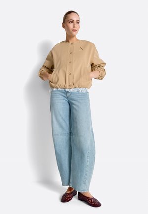 Moteris stovi su rankomis kišenėse, dėvi smėlio spalvos užsegamą švarkelį, šviesiai mėlynas platėjantis kelnes ir tamsius pintus batelius, priešais baltą foną.