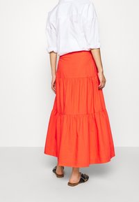 Henkilö, joka käyttää valkoista käärittyä paitaa, pitkää kirkkaan oranssia kerroksellista hametta ja mustia liukujalkineita, seisoo yksinkertaista valkoista taustaa vasten.