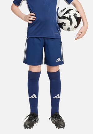 Uniforme de football navy à manches courtes, avec des rayures blanches contrastantes, associé à des chaussettes montantes assorties et des crampons noirs, tenant un ballon noir et blanc.