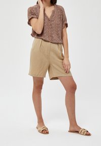 Donna che indossa pantaloni corti beige, una camicia a maniche corte a fantasia con bottoni e sandali di tela beige, in piedi con una mano vicino al viso.