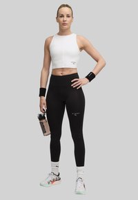 Witte geribbelde crop top en zwarte high-waisted leggings, met een heldere waterfles. Kenmerkt logo-accenten en sportschoenen met kleurrijke zolen.