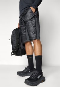 Shorts rembourrés noirs avec poches zippées, portés avec des chaussettes noires jusqu'aux genoux et des baskets HOKA. Un sac à dos noir est tenu dans la main droite.