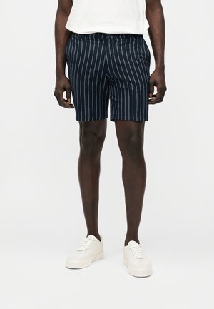 Άνδρας που φοράει σορτς ναυτικού μπλε με λευκές κάθετες ρίγες, λευκά αθλητικά παπούτσια και λευκό μπλουζάκι μαζεμένο μέσα, μπροστά σε απλό ανοιχτό φόντο.