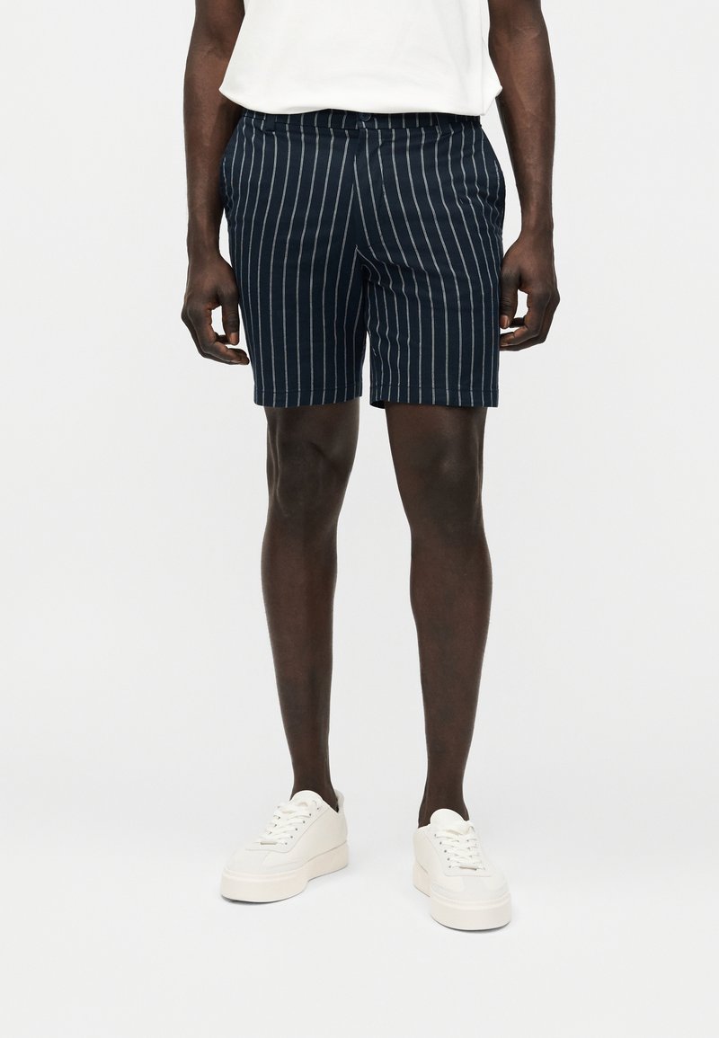 Άνδρας που φοράει σορτς ναυτικού μπλε με λευκές κάθετες ρίγες, λευκά αθλητικά παπούτσια και λευκό μπλουζάκι μαζεμένο μέσα, μπροστά σε απλό ανοιχτό φόντο.