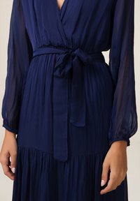 Robe bleu marine avec un design enveloppant, manches longues transparentes, une ceinture à nouer à la taille et une jupe plissée et fluide. Texture lisse, tissu léger.