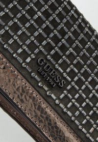 Musta tekstiilillä tehty Guess-laukku, jossa on hopeisia niittejä ruudukkomuodossa ja ruskeaa käärmeennahkaa korostavat reunat.