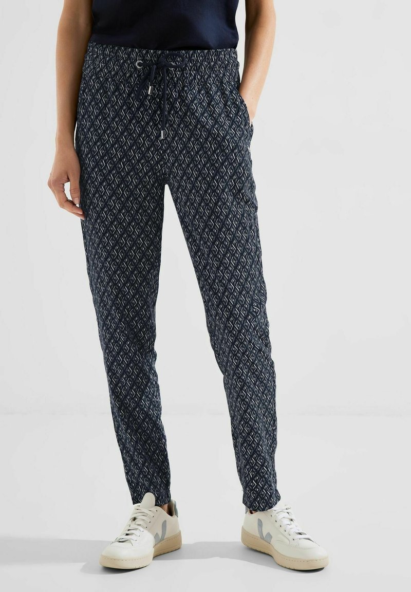 Femme portant un pantalon noir à motifs avec une taille à cordon et des baskets blanches, debout devant un fond clair uni.