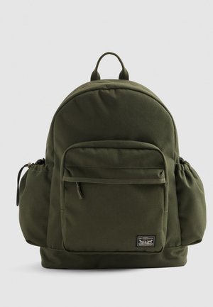 Zeleni tekstilni ruksak s zaobljenim oblikom, prednjim džepom na patentni zatvarač, bočnim mrežastim džepovima i gornjom ručkom. Ima brendiranu oznaku na prednjoj strani.