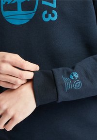 Sudadera navy con gráficos impresos en turquesa en el frente y la manga, con tejido texturizado y puños acanalados.
