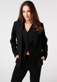 Zwarte op maat gemaakte blazer met piekboorden, twee voorzakken, gelaagd over een bijpassend mouwloos vest met knopen. Soepele stof, gestructureerde pasvorm.