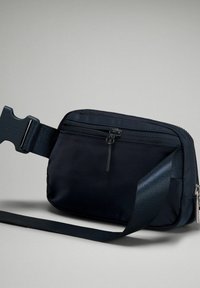 Černý nylonový páskový batoh se nastavitelným popruhem, hlavní kapsou na zip a plastovou sponou. Hladká textura s minimalistickým designem.