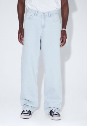 Mann trägt locker sitzende hellblaue Jeans, schwarz-weiße Sneakers und ein weißes Hemd, steht vor einem schlichten weißen Hintergrund.