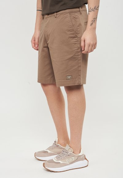 Pantaloni corti in cotone marrone con una tasca frontale, caratterizzati da un piccolo distintivo con logo. Abbinati a sneakers beige, che mostrano un design in suede testurizzato e mesh.