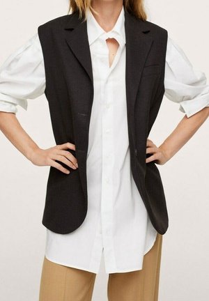 Femme portant une chemise blanche à manches longues boutonnée, un blazer noir sans manches et un pantalon beige, les mains sur les hanches.