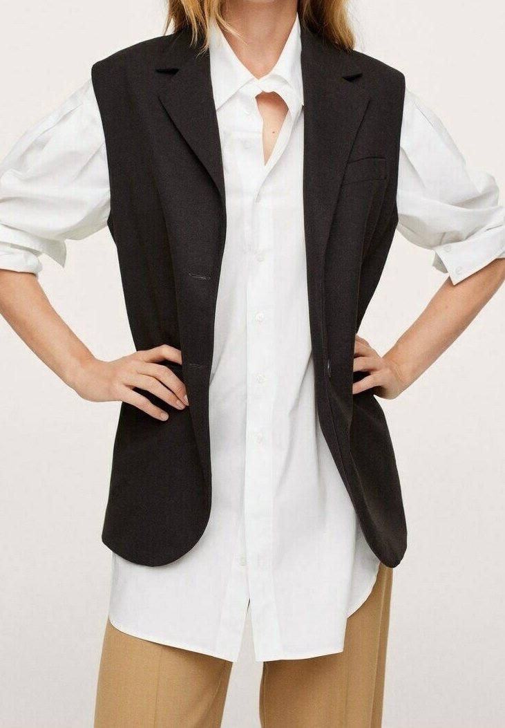 Femme portant une chemise blanche à manches longues boutonnée, un blazer noir sans manches et un pantalon beige, les mains sur les hanches.