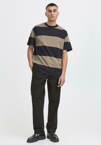 Camiseta a rayas en algodón de color azul marino oscuro y beige, con cuello redondo y corte holgado. Combinada con pantalones marrón oscuro y zapatos negros con acabado texturizado.