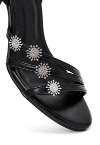 Sandale noire avec plusieurs fines lanières, comportant trois accents décoratifs en argent représentant des rayons de soleil, ornés de strass. Texture lisse, design à bout ouvert.
