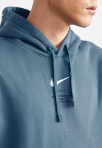 Sweatshirt com capuz em tecido teal com um logo da Nike branco no peito e um endereço impresso em roxo escuro. Possui cordões ajustáveis e uma textura suave.