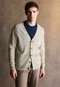 Light beige cardigan with a V-neck, five buttons, ribbed cuffs, and two front pockets, worn over a dark blue shirt. Smooth texture.