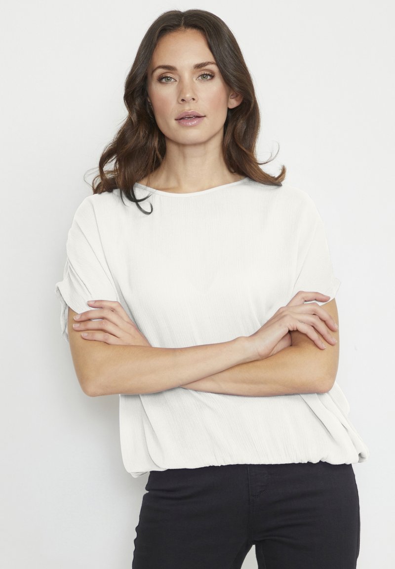 Haut blanc texturé avec une coupe ample et un large col, doté de manches courtes retroussées et d'un ourlet cintré, associé à un pantalon noir.