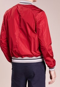 Uomo che indossa una giacca bomber di raso rossa con polsini e vita a strisce bianche e grigie, abbinata a pantaloni blu scuro, mostrato di spalle.