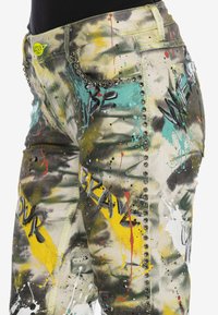 Gros plan sur un pantalon avec des éclaboussures de peinture colorée, des détails cloutés et des mots en style graffiti incluant "BRAVE" et "YOUR" sur le tissu.