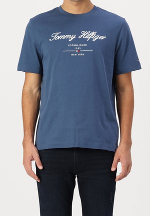 Man draagt een blauw Tommy Hilfiger T-shirt met korte mouwen, met een wit logo en de tekst "Established 1985 New York" aan de voorkant, gecombineerd met een donkere broek.