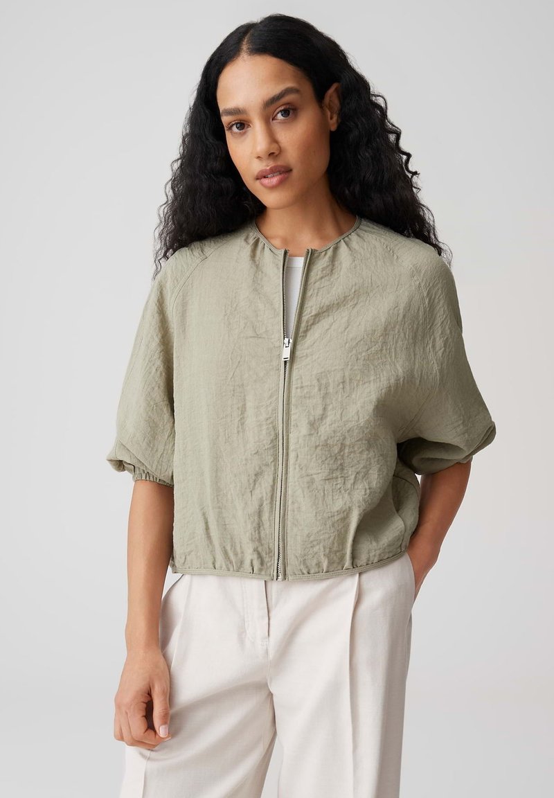 Femme aux longs cheveux bouclés portant une veste zippée olive clair à manches courtes et un pantalon blanc taille haute, debout devant un fond uni.