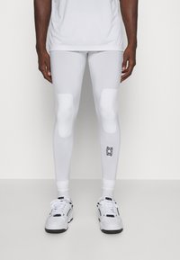 Leggings atletici bianchi con accenti imbottiti sulle ginocchia, texture liscia e un piccolo logo nero vicino alla caviglia. Abbinati a sneakers bianche.