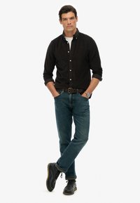 Camisa negra de botones, camiseta blanca de cuello redondo debajo, jeans de mezclilla azul, botas negras. El modelo está de pie con los brazos cruzados, mostrando un look casual.